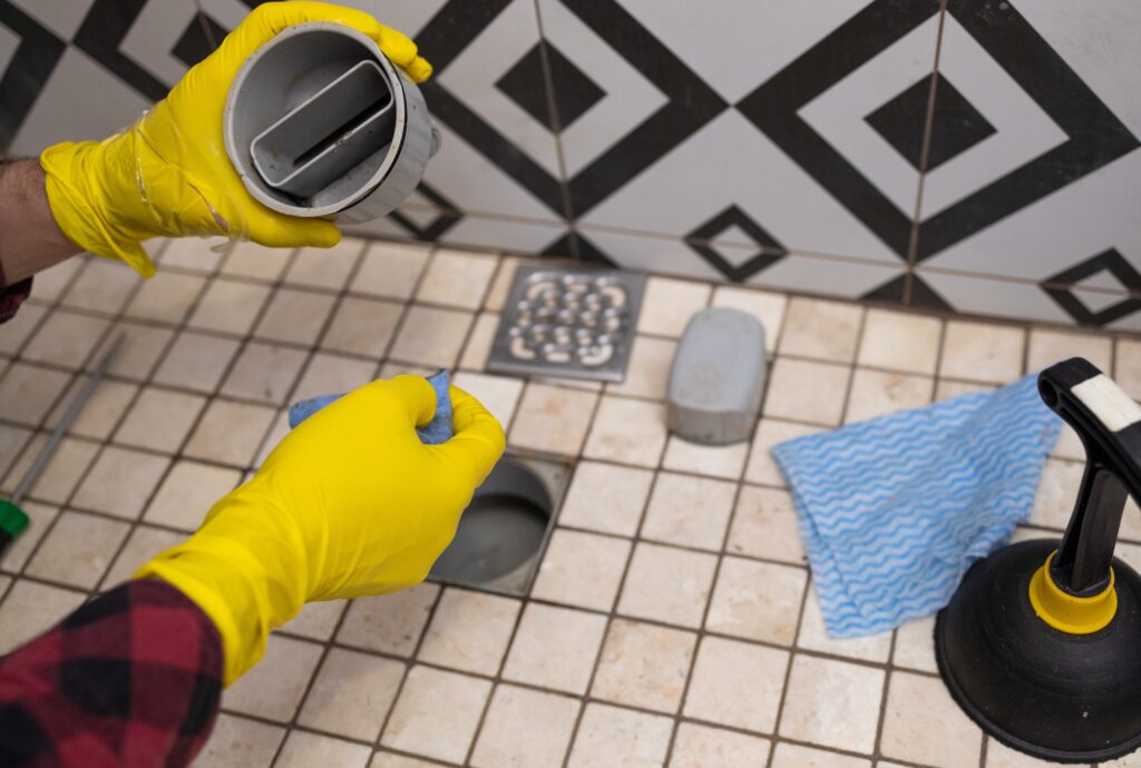 Hands in yellow gloves removing a drain cover to unblock shower drain and clear water blockage on a tiled bathroom floor.